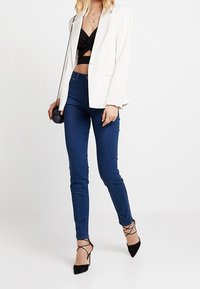 Blazer blanco texturizado sobre un top corto negro, combinado con jeans ajustados de mezclilla azul oscuro y tacones altos negros con tiras.