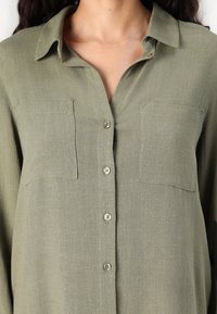 Chemise boutonnée vert olive en tissu léger, à manches longues, avec deux poches poitrine et un col, présentant une texture subtile.