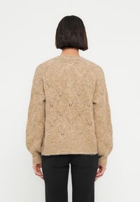 Γυναίκα που φαίνεται από πίσω φοράει μπεζ πλεκτό πουλόβερ με διαμάντι σχέδια και μαύρο παντελόνι, σε απλό λευκό φόντο.