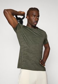 Olive green camouflage t-shirt with short sleeves, round neckline, and a light texture, worn by a person holding a kettlebell overhead.