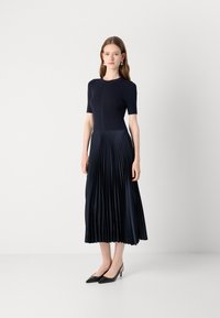Navy blue dress with a fitted ribbed bodice and pleated skirt. Short sleeves, knee-length with a smooth texture. Black pointed heels.