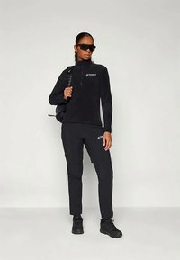 Schwarzes Outdoor-Outfit, bestehend aus einem Fleece-Oberteil mit Reißverschluss und enganliegenden Hosen, beide mit einem Logo, kombiniert mit schwarzen Sneakers und Sonnenbrille.