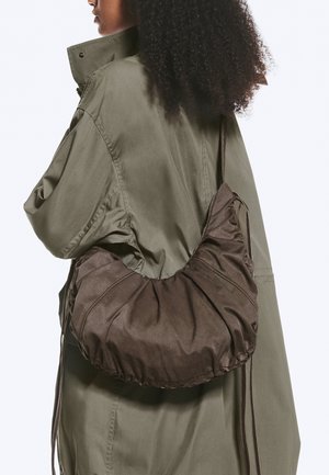 Personne portant une veste vert olive, portant un grand sac à bandoulière marron foncé froncé avec une seule bandoulière dans le dos.