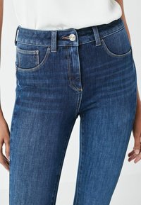 Jeans de mezclilla azul oscuro con talle alto, que cuentan con un cierre de botón frontal, cinco bolsillos y detalles de costura en contraste.