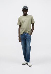 T-shirt di cotone verde oliva con piccolo logo sul petto, abbinata a jeans denim azzurri chiari e cappellino grigio. Scarpe da ginnastica bianche e grigie completano il look.