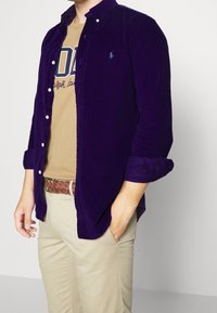 Violetti corduroy-nappipaita, jossa on kaulus, käännetyt hihat ja hillitty logo rinnassa, yhdistettynä ruskeaan t-paitaan ja beigenvärisiin housuihin.