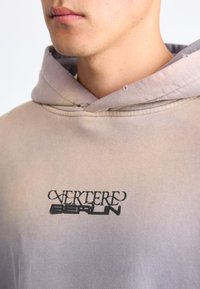 Hooded sweatshirt in a gradient of beige to lavender. Features a black logo "VERTERE BERLIN" on the chest. Made of soft cotton fabric.
