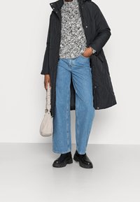 Černý dlouhý prošívaný kabát, vzorovaný pletený svetr, světle modré džíny s širokými nohavicemi, černé robustní boty a světle béžová kabelka přes rameno.