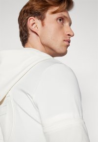 Hvit hettegenser med teksturert design på skuldrene og ryggen. Har snøring på hetten og glatt stoff. Nærbilde av nakke og skulder.