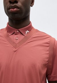 Κοντινό πλάνο ενός ατόμου που φοράει μπλούζα dusty rose με V-λαιμόκοψη πάνω από πουκάμισο με γιακά, με λευκό λογότυπο στον γιακά.