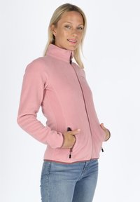 Giacca di pile rosa con colletto alto, chiusura con zipper frontale e tasche laterali. Presenta un design aderente con cuciture a contrasto e texture.