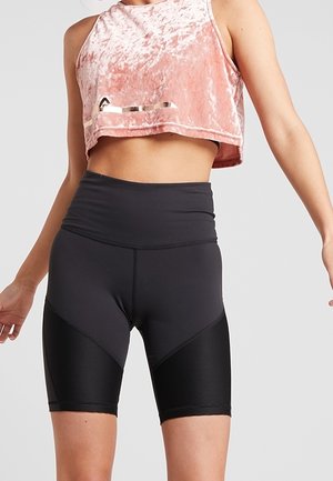 Femme portant un débardeur court en velours rose et un short de cycliste noir taille haute, debout devant un fond uni.