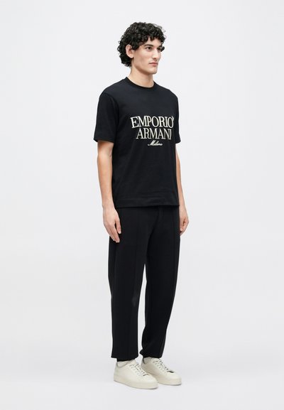 Μαύρο βαμβακερό μπλουζάκι με λευκό λογότυπο "Emporio Armani", συνδυασμένο με φαρδιά μαύρη παντελόνια και εκρού αθλητικά παπούτσια, σε απλό φόντο.