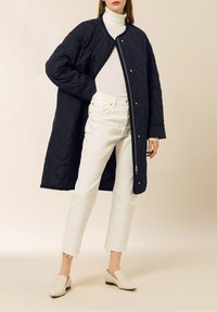 Abrigo largo acolchado azul marino con cuello redondo, combinado con jeans blancos de pierna recta y zapatos beige sin cordones, sobre un fondo neutro.