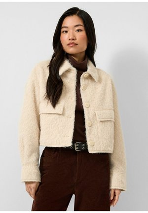 Femme portant une veste crème duveteuse boutonnée sur un col roulé marron et un pantalon en velours côtelé marron avec une ceinture noire sur un fond uni.