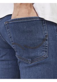 Hand inserted into back pocket of blue denim jeans worn with a white shirt.