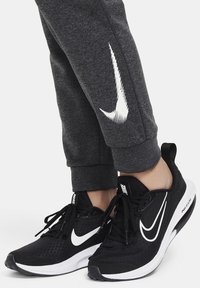 Zwarte Nike sneakers hebben een bovenwerk van mesh, een witte swoosh en een gewatteerde kraag. Grijze joggers met een witte swoosh-logo op het linkerbeen.