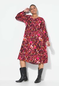 Langärmliges A-Linien-Kleid mit einem lebhaften Muster in Pink, Rot und Schwarz, das wirbelt. Kombiniert mit schwarzen Cowboystiefeln.