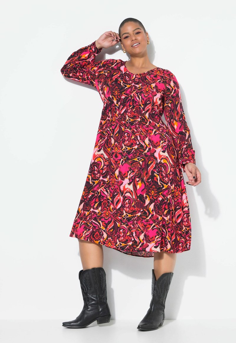 Langärmliges A-Linien-Kleid mit einem lebhaften Muster in Pink, Rot und Schwarz, das wirbelt. Kombiniert mit schwarzen Cowboystiefeln.