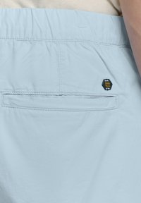 Lichtblauwe shorts met een gladde textuur, voorzien van een achterzak en een klein zwart en geel hexagonaal logo aan de zijkant.
