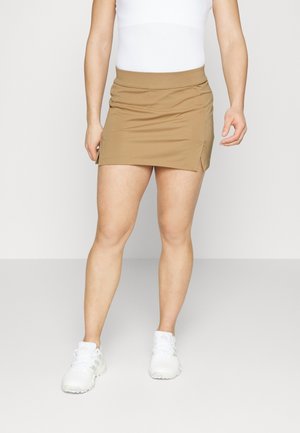 Person wearing a white sleeveless top, tan mini skirt with side slit, and white athletic shoes, standing against a plain background.