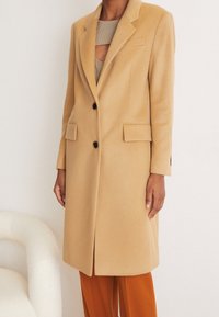 Cappotto in misto cammello con un design strutturato, davanti a un bottone singolo, rever a taglio e due tasche frontali. Bottoni neri aggiungono contrasto.