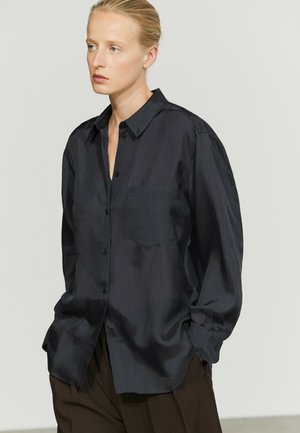 Woman with blonde hair tied back wearing a loose black button-up satin shirt and brown pants, looking to the side with hands in pockets.