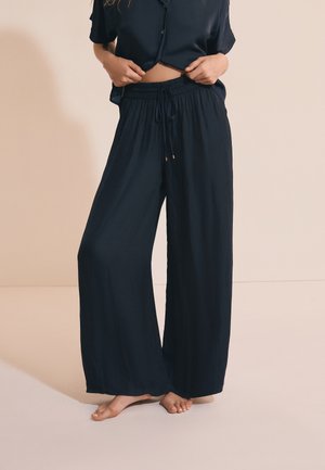 Frau trägt lockere schwarze weite Hosen mit Kordelzug in der Taille, barfuß, und ein passendes schwarzes kurzärmeliges Hemd mit Knöpfen, leicht angehoben.