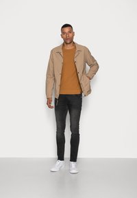Veste légère beige avec des boutons-pression, pull orange, jeans slim noirs et baskets blanches. Modèle posé contre un fond uni.