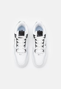 White sneakers with black accents, featuring a smooth leather upper, textured lining, laces, and a signature logo on the tongue.