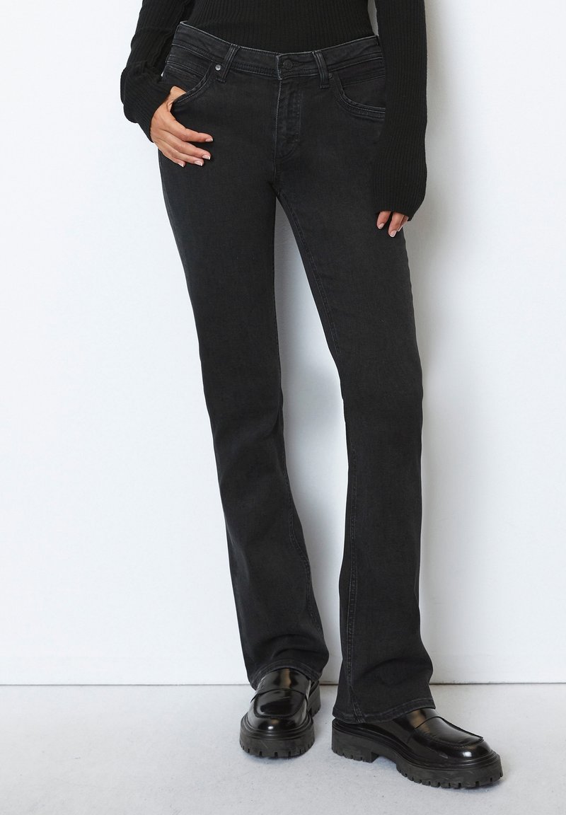 Jean flare en denim noir avec un design classique à cinq poches, taille mi-haute et tissu texturé, associé à des chaussures noires imposantes.