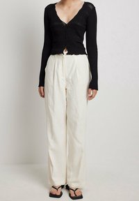 Schwarzes, geripptes Langarmoberteil mit V-Ausschnitt und Frontknöpfen, kombiniert mit lockeren cremefarbenen Leinenhosen und schwarzen Riemchensandalen.
