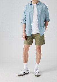 Mies, päällään vaaleansininen nappipaita valkoisen t-paidan päällä, oliivinvihreät shortsit, valkoiset sukat ja valkoiset tennarit, seisoo yksi käsi taskussa.