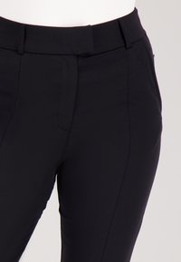 Close-up van zwarte, getailleerde broek met riemlussen en een verborgen sluiting aan de voorkant op een witte achtergrond.
