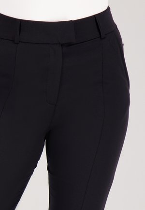 Close-up van zwarte, getailleerde broek met riemlussen en een verborgen sluiting aan de voorkant op een witte achtergrond.