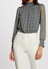 Durchsichtige, gemusterte Bluse mit hohem Ausschnitt und langen Ärmeln, die komplizierte geometrische Designs in Blau- und Weißtönen zeigt.