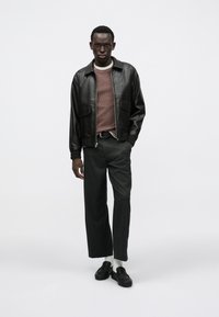 Casaco de couro preto com dois bolsos frontais, camisola canelada bordô, calças cinzentas e mocassins pretos. Modelo está em pé contra um fundo branco.