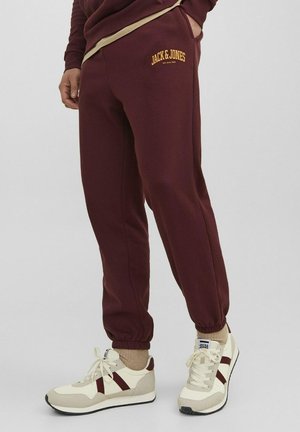 Jogging in borgogna con polsini elastici, caratterizzati da un logo giallo. Abbinati a sneakers crema con accenti marroni e un calzino beige testurizzato.