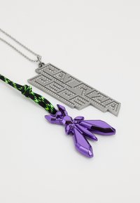 Fém medál gravírozott szöveggel, tiszta kristályokkal díszítve, egy lila akril charm-mal, fekete és zöld fonott zsinóron.