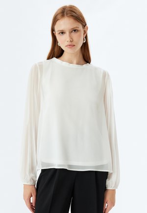 Blusa bianca trasparente con maniche lunghe e gonfie, scollatura a barca e tessuto leggermente trasparente. Indossata sopra pantaloni neri.