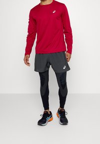 T-shirt de sport rouge à manches longues avec logo, short gris, leggings noirs avec accents texturés, et chaussures de course noires avec des détails orange et bleu.