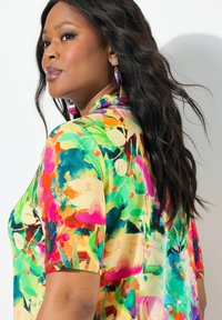 Barevná blůza s krátkým rukávem, která se pyšní živými květinovými vzory v zelených, růžových a žlutých tónech. Hladký materiál s uvolněným střihem a límečkem.