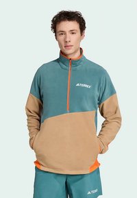 Polárdzseki fél cipzáras kialakítással; kék felső rész, barna alsó rész, élénk narancssárga díszítések és 'TERREX' logó a mellkason.