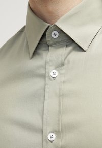 Hellgrünes Hemd mit klassischem Kragen, glattem Stoff und weißen Kunststoffknöpfen auf der Vorderseite. Die Nähte sind ordentlich und schlicht.