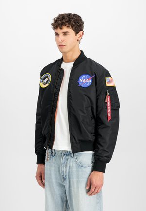 Young man with curly hair wearing black bomber jacket with NASA and American flag patches, white shirt, and light blue jeans.