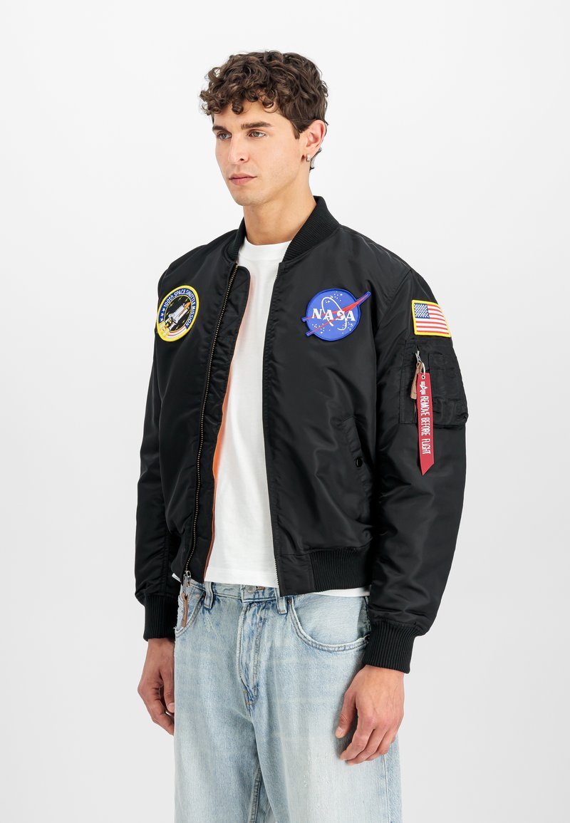Giovane uomo con capelli ricci che indossa una giacca bomber nera con toppe NASA e bandiera americana, camicia bianca e jeans azzurri.