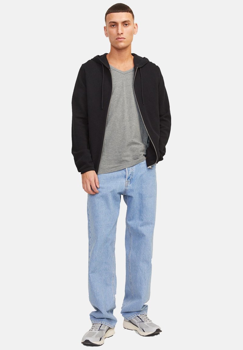 Jeune homme debout portant un sweat à capuche zippé noir, un T-shirt gris à col en V, un jean bleu clair et des baskets grises, sur un fond uni.