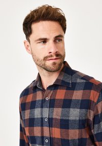 Mann mit kurzen braunen Haaren und Bart, der ein gestreiftes, blaues, braunes und graues Button-up-Hemd trägt und nach links schaut.