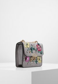 Borsa a tracolla in suede grigio con catena dorata, ricami floreali e decorazioni di gioielli su una superficie di esposizione bianca.