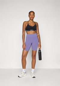 Brassière de sport noire avec logo blanc, associée à un short violet taille haute. Le mannequin tient une bouteille d'eau noire, portant des baskets et des chaussettes blanches.